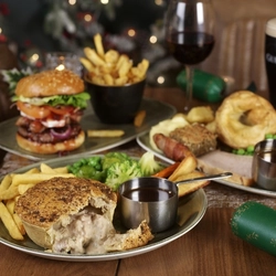 A close up view of a Turkey and Ham Pie served on a plate with chips, vegetables and a small pot of gravy. The plate sits on a wooden table along with a plated roast dinner, a Festive Burger served on another plate with a bowl of fries, and glasses of red wine and Guinness.