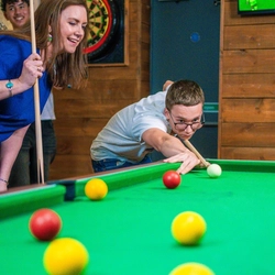 A person leans over a pool table, aiming their cue toward the cue ball while preparing to take a shot. Another person stands close beside them, holding a cue stick and watching the play. The setting is a wood‑paneled games room with a dartboard mounted on the wall behind them and a TV screen in the corner displaying a sports match. The scene captures a focused, social moment during a game of pool.