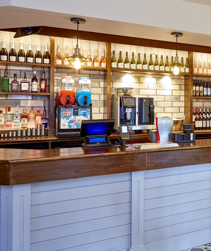 A close up view of the bar inside The Flying Horse in Heald Green. Behind the bar are bottles of wine and spirits on wooden shelves, as well as a slushie machine.