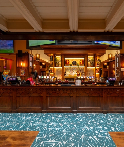 The wood panelled bar inside the Old School House, with TVs on the wall above it, and bottles of alcoholic spirits on illuminated shelves behind the counter.