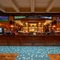 The wood panelled bar inside the Old School House, with TVs on the wall above it, and bottles of alcoholic spirits on illuminated shelves behind the counter.