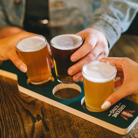 A Beer Flight Paddle is on an indoor wooden table.  There are the hands of three people seated at the table, each taking a mini pint glass of beer from it.