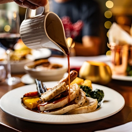 A lifestyle image showing a jug of gravy being poured over a plated Christmas/Festive main dish sat on a table within the interior restaurant seating area.