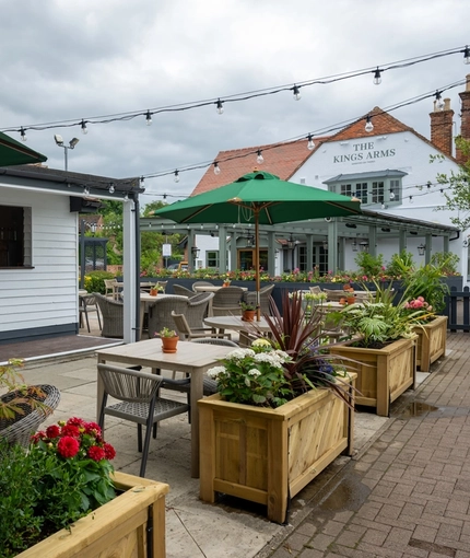 The exterior bar and beer garden seating area at The Kings Arms in Sandford-On-Thames, with wooden planter boxes, shade umbrellas on some of the tables, and string lights overhead.