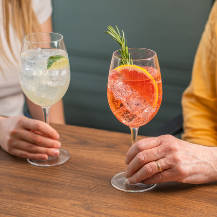 A Hugo Spritz cocktail and a Lillet Spritz cocktail, each served with ice, in a wine glass which stands on a wooden table. The Hugo Spritz is garnished with a slice of lime, the Lillet Spritz is garnished with a sprig of rosemary and a slice of grapefruit, and two people are sitting at the table, each holding one of the glasses.