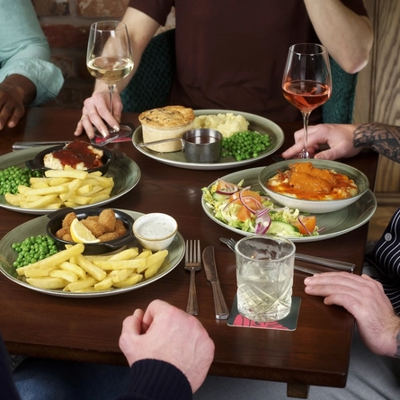 A round table filled with main dishes, including scampi and chips and a pie with mash and peas. Several drinks are placed around the table. The hands and arms of multiple people seated together are visible as they reach toward their food and raise their drinks.
