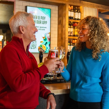 A male and female stood at the bar holding drinks in a cheers position in a Flaming Grill venue. 
