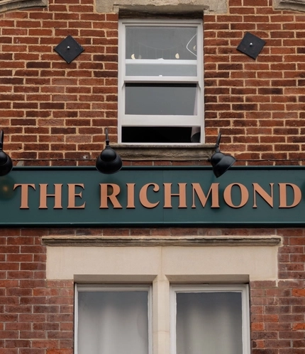 A close up view of the exterior facade of The Richmond Arms, with a green and gold pub sign on the brick wall in between two upper windows.