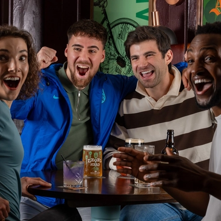 Four people sit closely together around a small pub table with drinks on it. All four individuals have their mouths open, as if cheering and are looking toward something out of frame. One person has an arm raised with a bent elbow, another has an arm around the shoulders of the person next to them, and the table holds glasses, a beer can, and a bottle.