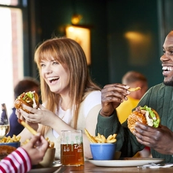 A mixed group of people seated at an indoor wooden table, enjoying burgers and conversation.