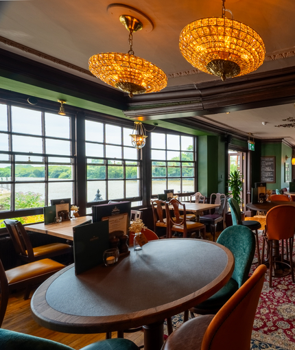 The interior restaurant seating area at The London Apprentice, with wooden flooring, two jewelled ceiling lights, windows overlooking the River Thames, a fireplace, and decorative plaster on the ceiling.