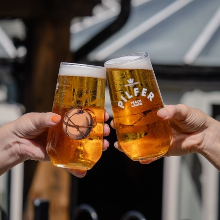 A close up lifestyle image showing 2 people holding pints of Pilfer Peach Lager and celebrating with a cheers.