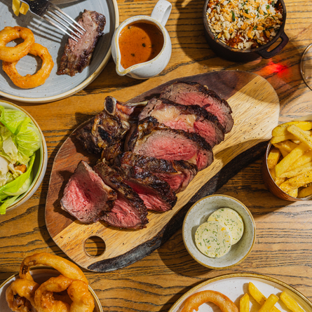 An advertising image showing a boarded Ribeye Sharer dish including onions rings, chips, salad and sauce sat on a table within the interior restaurant seating area at The Four Oaks.