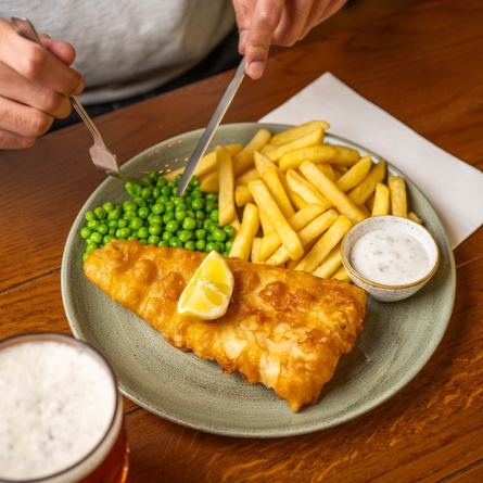 Fish & Chips served with peas, lemon, and a small pot of dipping sauce, on a plate which sits on a wooden restaurant table. A glass of beer and a napkin sit on either side of the plate, and a person sitting at the table is picking up peas with their knife and fork.