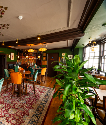 An interior restaurant seating area at The London Apprentice, with a small bar, two fireplaces, framed artwork on the walls, and painted decorative plaster on the ceiling in the shape of red grapes on vines.