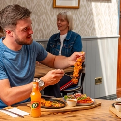 Two people sitting at a wooden restaurant table. One person has a plate of steak and chips on the table in front of them and takes a sip of Heineken 0.0% alcohol beer from the bottle; the other person is holding a skewer of chicken and is using a fork to remove a piece. A bottle of J2O soft drink sits on the table next to them.