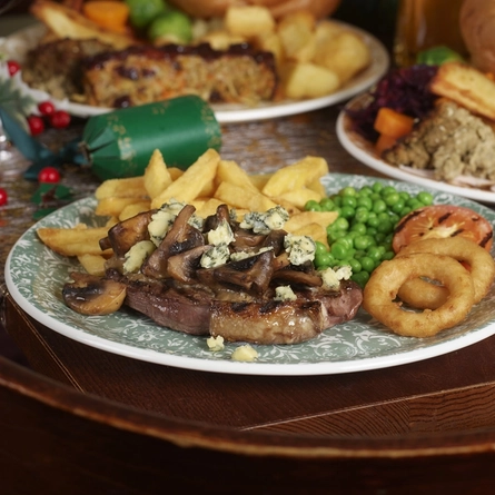 An advertising image focusing on a Christmas Day Stilton Steak Carvery dish with various plated Festive Main dishes in the background and a selection of drinks available at a Pub & Carvery venue.
