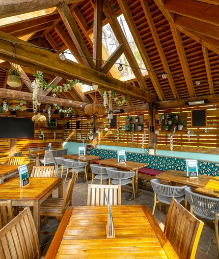 The exterior under cover seating area at The Walkford, with string lights and flowers hanging from the vaulted ceiling, potted plants and chalkboards on the wooden walls, and padded bench seats along one wall.