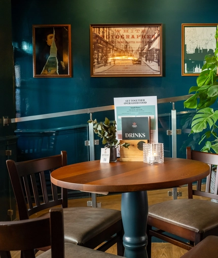 The interior restaurant seating area of the Cock & Pye, with wooden furniture, a large potted plant, and framed artwork on the wall.