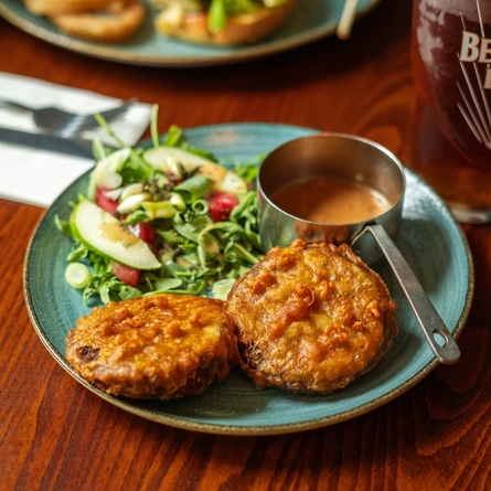 Haggis fritters served on a plate with salad and a pot of sauce. The plate sits on a wooden table next to a glass of beer, cutlery, a napkin, and another plate of food.