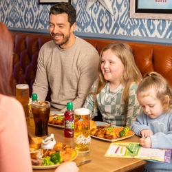People sit on a leather bench around a wooden table in a pub, eating a roast meal with vegetables and potatoes. Drinks including pints of lager and soft drinks are on the table, and a child colours in a book beside a plate of food.