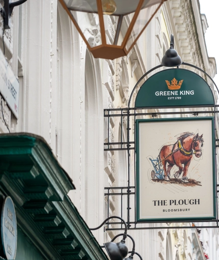 A close up view of the pub sign hanging on the exterior wall of The Plough. The sign features the Greene King logo, and the words "The Plough, Bloomsbury" below an illustration of a horse pulling an old fashioned plough.