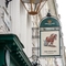A close up view of the pub sign hanging on the exterior wall of The Plough. The sign features the Greene King logo, and the words "The Plough, Bloomsbury" below an illustration of a horse pulling an old fashioned plough.