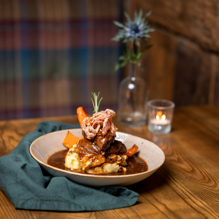 A dish of mash, gravy, and a yorkshire pudding. Next to a blue napkin, in front of tartan curtains with a thistle in a vase behind. 