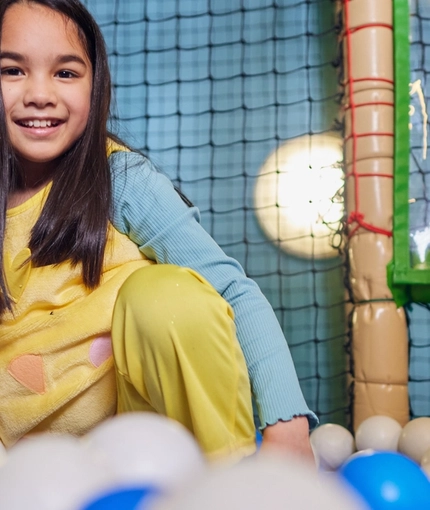A child smiling at the camera in a ball pit in an indoor play area.
