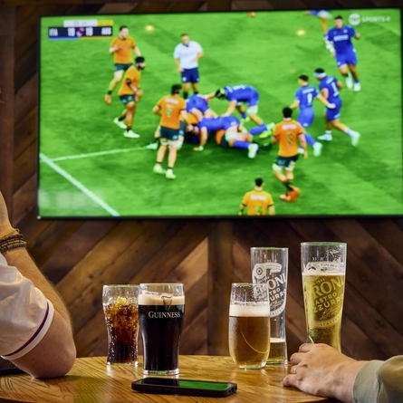 Two people sitting at a table in a pub, watching sports on the TV on the wall in front of them. A round of drinks and a mobile phone sit on the table between them.