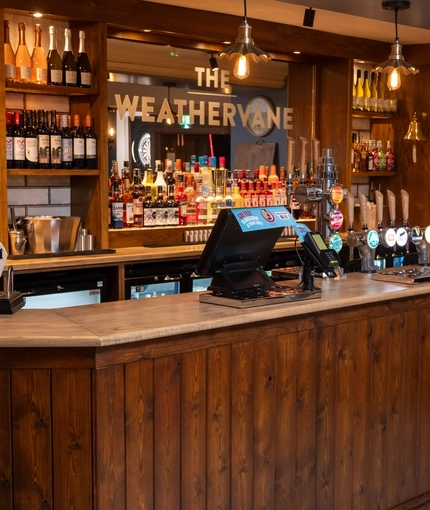 A close up view of the wood panelled bar inside The Weathervane, with bottles of wine and alcoholic spirits on wooden shelves behind the counter.