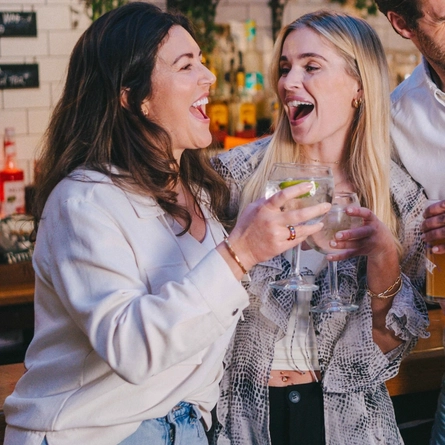 Four people standing beside the bar in a pub with their arms around each other. Two of them are holding glasses of beer, and the other two are holding cocktails.