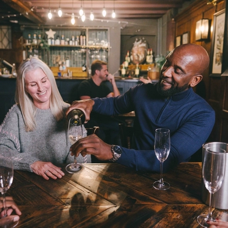 A lifestyle image focusing on 2 customers sat at a table with friends while one of the customers pours out glasses of sparkling white wine to celebrate Mothers Day within the interior seating area at a Heritage venue.