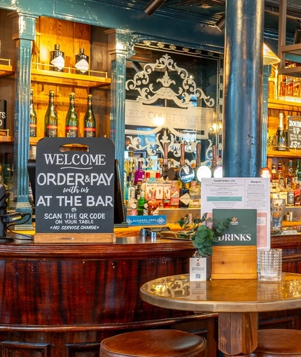 The wood panelled bar inside The Earls Court Tavern, with bar stool seating, and wine glasses hanging in racks above the counter.