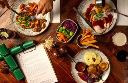 An overhead shot of a range of plated Christmas mains on a festive decorated wooden table, with hands of a person seated around the table holding a knife and fork.