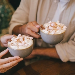 Two hands holding cups of hot chocolate topped with cream, a chocolate flake and mini marshmallows..