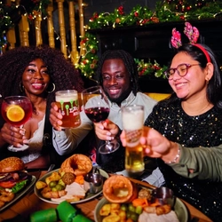 A group of people celebrating a festive Cheers while enjoying various main meals and drinks within the interior restaurant and seating area of Greene King Pub venue.