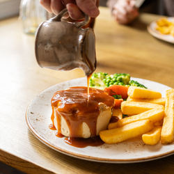 A close up image showing a person pouring gravy over a plated Mushroom & Ale Pie dish sat on a table within the interior restaurant seating area at a Chef & Brewer venue.
