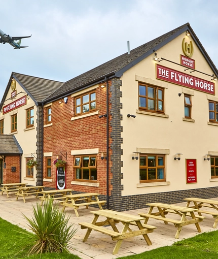 The exterior facade, signage and beer garden seating area with wooden picnic tables, at The Flying Horse in Heald Green.