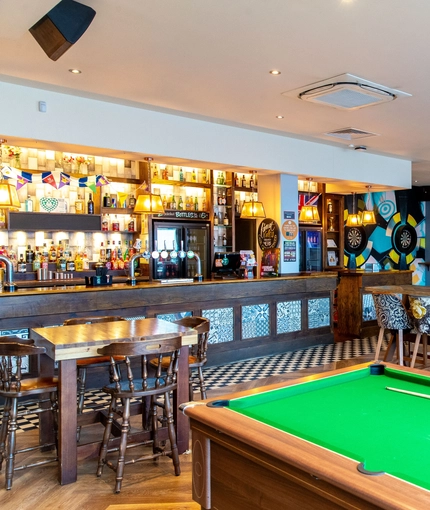 Interior dining area of a pub with a bar, a TV, a fireplace, a pool table and a few dartboards.