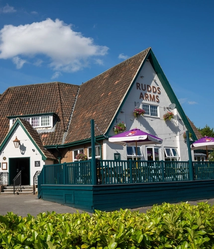 The exterior facade, signage, and seating area of the Rudds Arms in Marton-In-Cleveland; with an accessibility ramp leading up to the door; and picnic tables, shade umbrellas, and string lights on a raised deck area.
