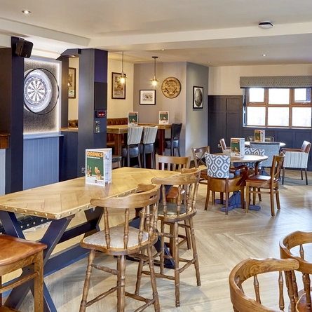 The interior restaurant seating area of The Flying Horse in Heald Green, with wooden tables, upholstered chairs, a TV on the wall and a dartboard.