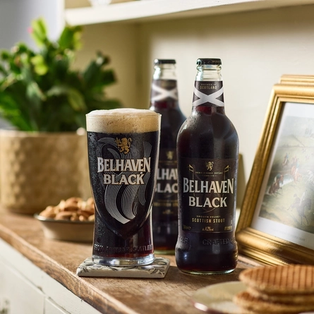 An advertising image of a pint and two bottles of Belhaven Black sat on a sideboard within a living room setting.