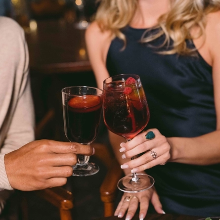 Two people sitting at a wooden restaurant table, clinking together a glass of mulled wine and a cocktail served in a wine glass.