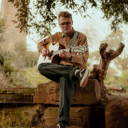 A image of a performer sat on a rock outside playing the guitar, used to advertise 'whats on' in Crafted venues.