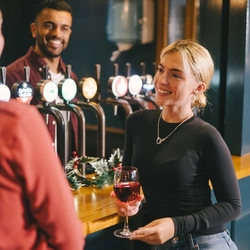 An image of a person stood behind the interior bar and 2 customers stood in front of the bar with drinks.