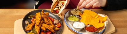 A board holding a skillet of chicken and a plate of dips, nachos and a wrap on a wooden table.  A person seated at the table is holding a fajita.