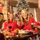 A group enjoys a Christmas meal in a pub decorated with a snow-covered Christmas tree and festive red and gold baubles. One guest wearing a paper crown holds up a Brussels sprout on a fork while laughing with others around the table. Christmas crackers, drinks and a traditional festive dinner are spread across the table, creating a lively and sociable holiday dining scene.