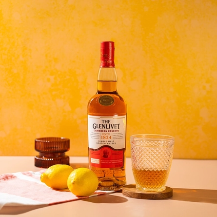 A bottle of Glenlivet Caribbean Reserve Single Malt Scotch Whisky, standing on a table in front of a yellow background. A glass bowl, a napkin, and two lemons also sit on the table; as well as a lowball glass half filled with the whisky which stands on a wooden coaster.