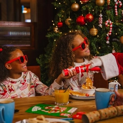 A festive breakfast table set beside a decorated Christmas tree adorned with red and gold baubles, candy cane ornaments and warm fairy lights. Two children wearing Christmas-print pyjamas and red novelty sunglasses sit at the table, with one holding a red Christmas cracker being handed over by a person dressed as Santa Claus. Breakfast dishes, mugs, juice cartons, a glass of orange juice, Christmas crackers and a colouring activity sheet are arranged on the wooden table, while other diners sit nearby in the Christmas-themed restaurant setting.